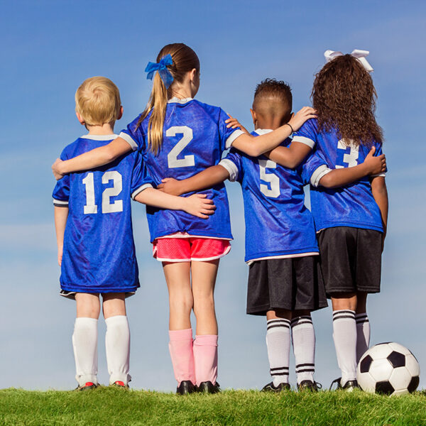 Four children in blue soccer jerseys with numbers 12, 2, 5, and 34 stand arm-in-arm on grass, facing away from the camera. A soccer ball is on the ground beside them.
