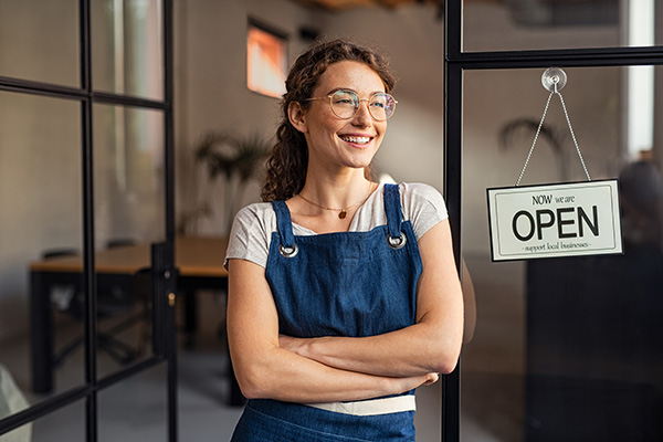 Smiling woman with glasses stands by an open sign in a modern room, wearing a blue apron over a white shirt.