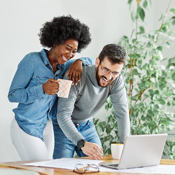 Two people smiling while looking at a laptop, one holding a coffee mug, in a bright room with plants.
