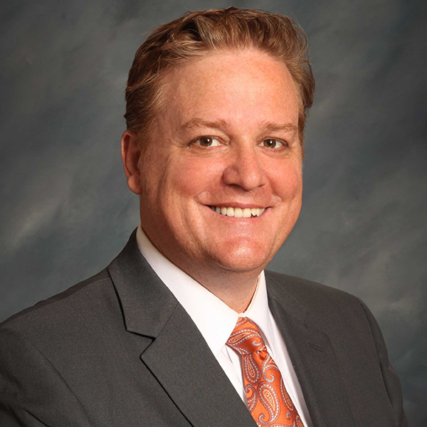A man wearing a dark suit, white shirt, and patterned orange tie is smiling against a neutral gray background.