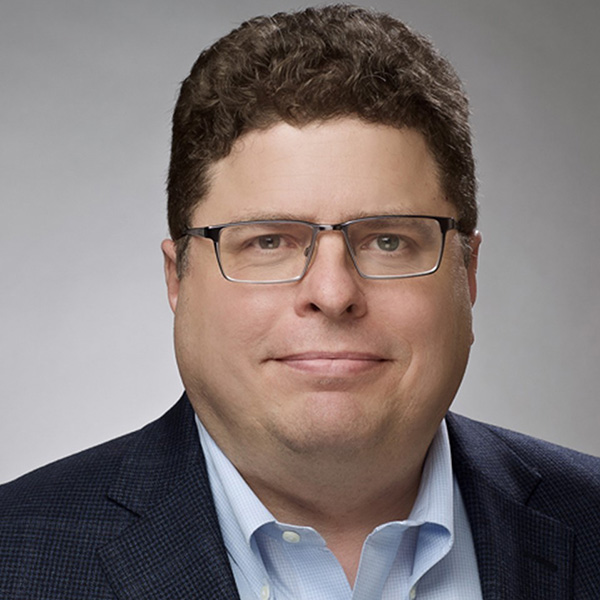 Man with glasses and curly hair in a suit jacket and shirt, smiling softly against a gray background.