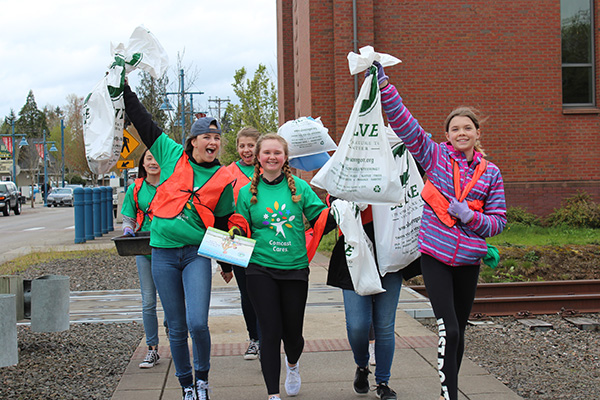 Group of people wearing green shirts and orange vests holding up garbage bags while walking on a sidewalk, participating in a clean-up event.