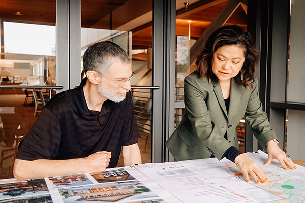 Two people discuss architectural plans at a table. The man is seated, and the woman points at documents. They are in a modern room with large glass doors.