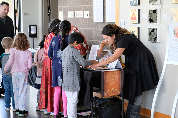 A woman assists a line of children in colorful clothing at a tabletop activity in a gallery setting with art displayed on the walls.