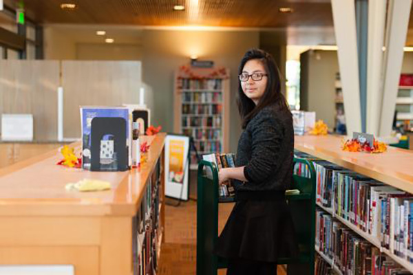 A person posing for the camera in a library with a cart of books next to her.