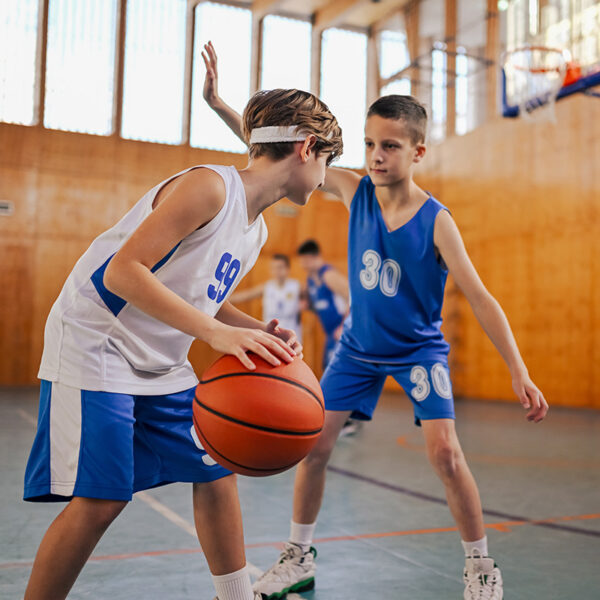 Two young boys playing basketball indoors. One boy, in a white jersey, is dribbling the ball while being closely guarded by another in a blue jersey.