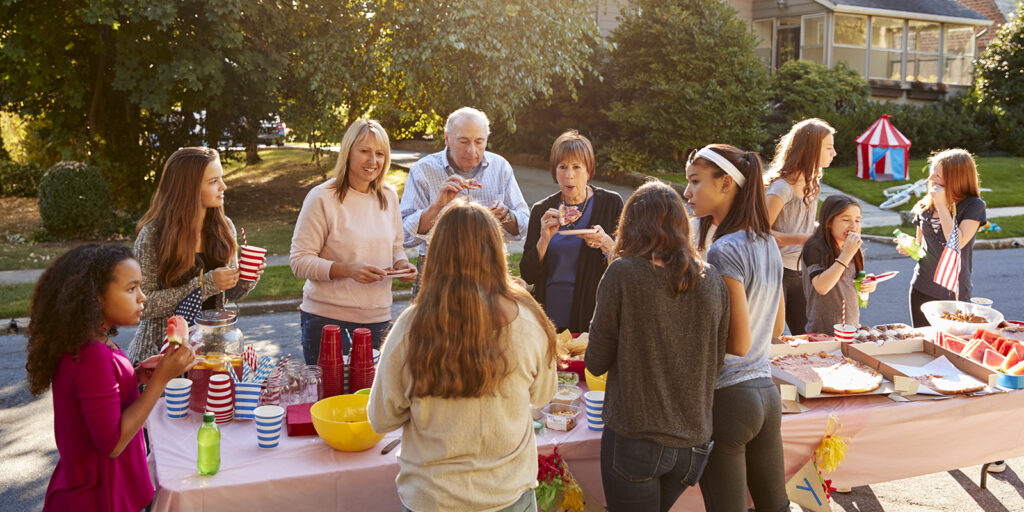 A group of people of various ages gathers around an outdoor table with food and drinks. They are eating and socializing in a suburban setting.