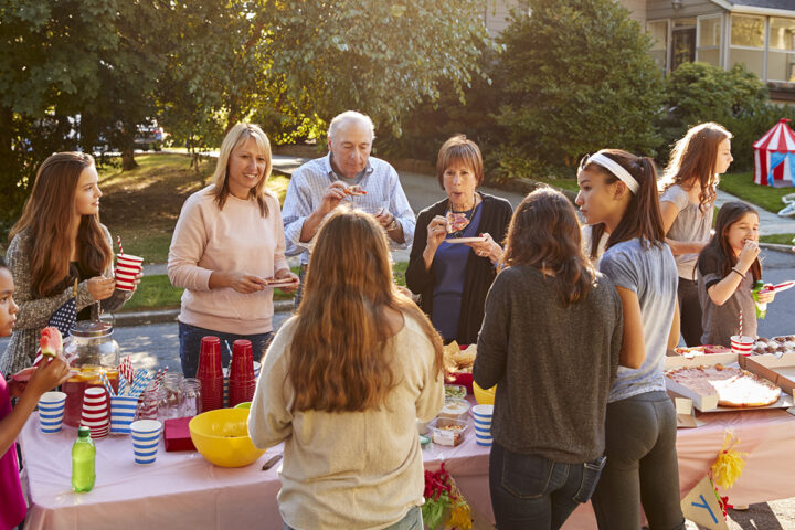 A group of people of various ages gathers around an outdoor table with food and drinks. They are eating and socializing in a suburban setting.