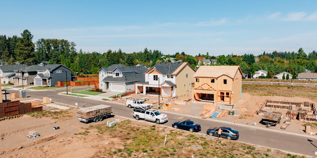 Residential neighborhood with houses under construction, some completed. Trucks and cars parked on the street. Wood materials and construction equipment visible on site.
