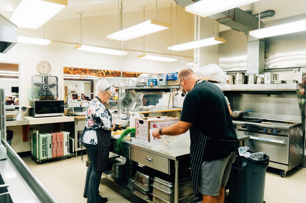 Two people in a commercial kitchen preparing food. They are wearing aprons and hairnets, and working around a metal prep table with boxes and vegetables.