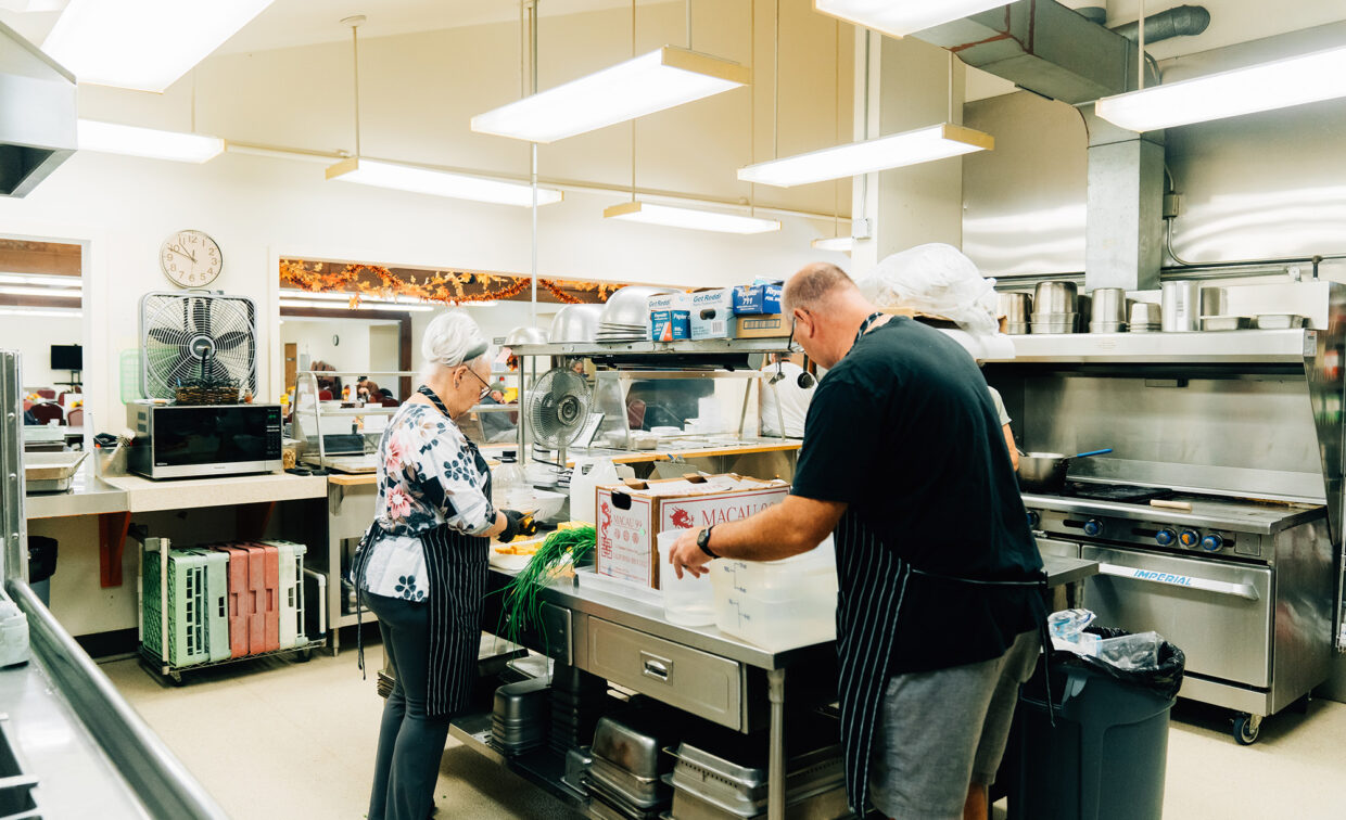 Two people in a commercial kitchen preparing food. They are wearing aprons and hairnets, and working around a metal prep table with boxes and vegetables.