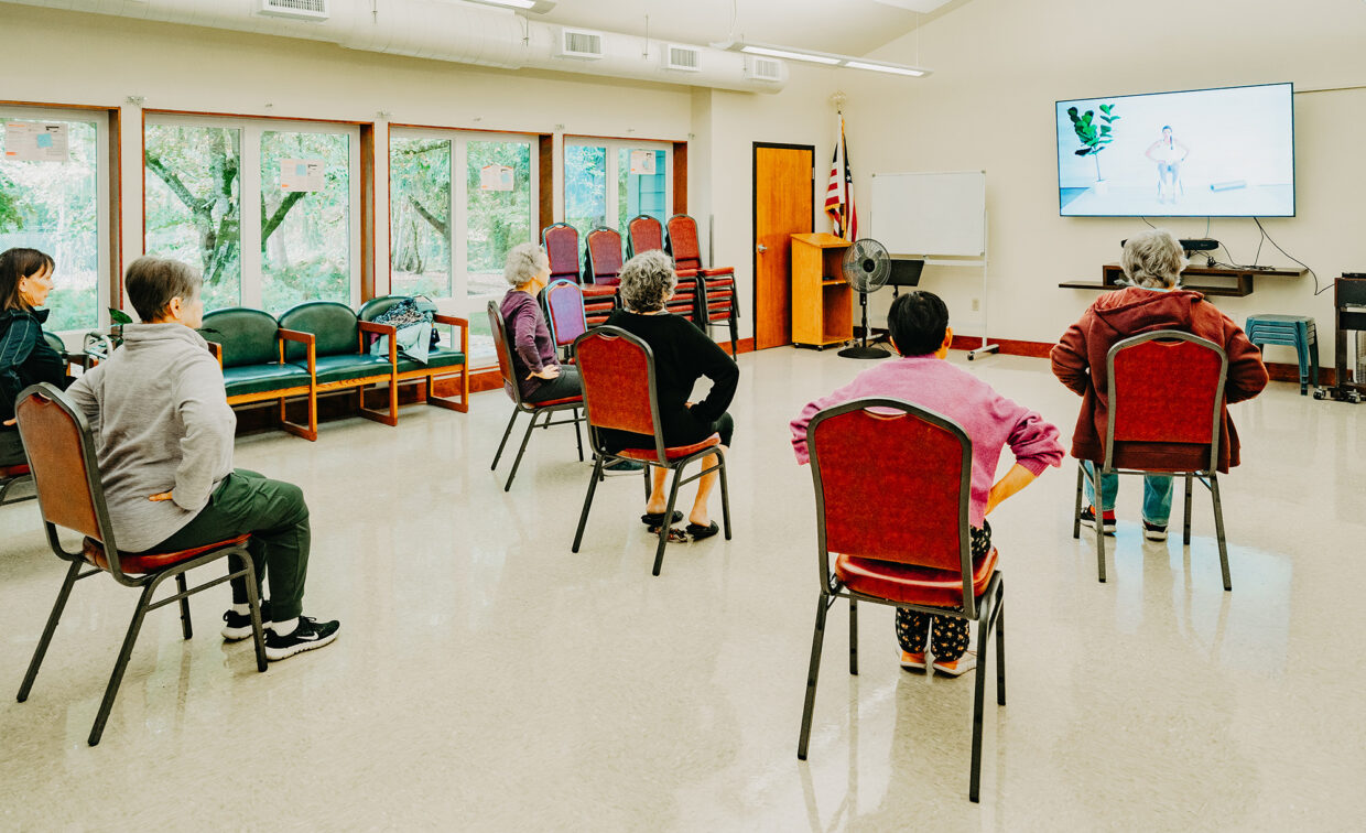 A group of people sitting on chairs in a community center, following a fitness routine displayed on a large TV screen.