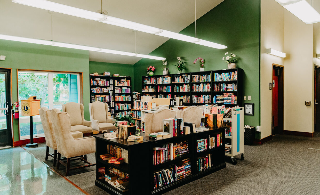 A library corner with shelves filled with books, armchairs, a book cart, and green walls.
