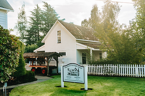 White house with a gabled roof, surrounded by a white picket fence and trees. A sign in front reads 