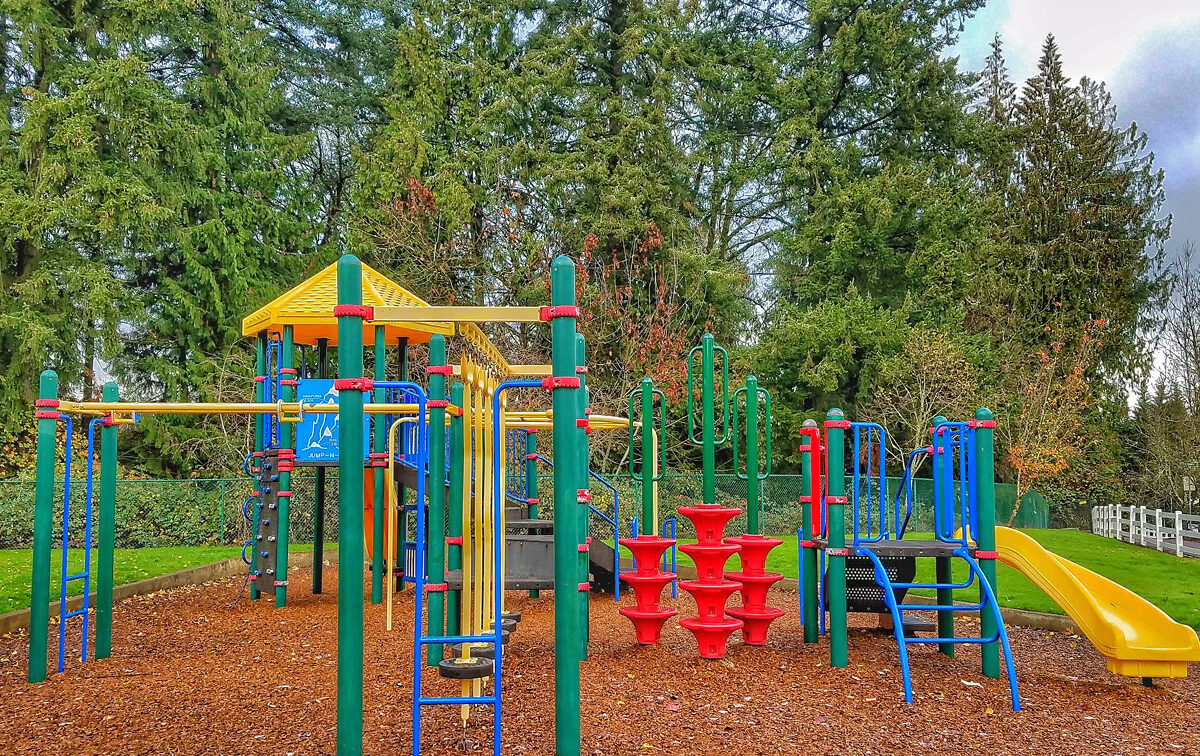 Colorful playground with various equipment, including slides, climbing structures, and monkey bars, set on a woodchip surface with tall trees in the background.