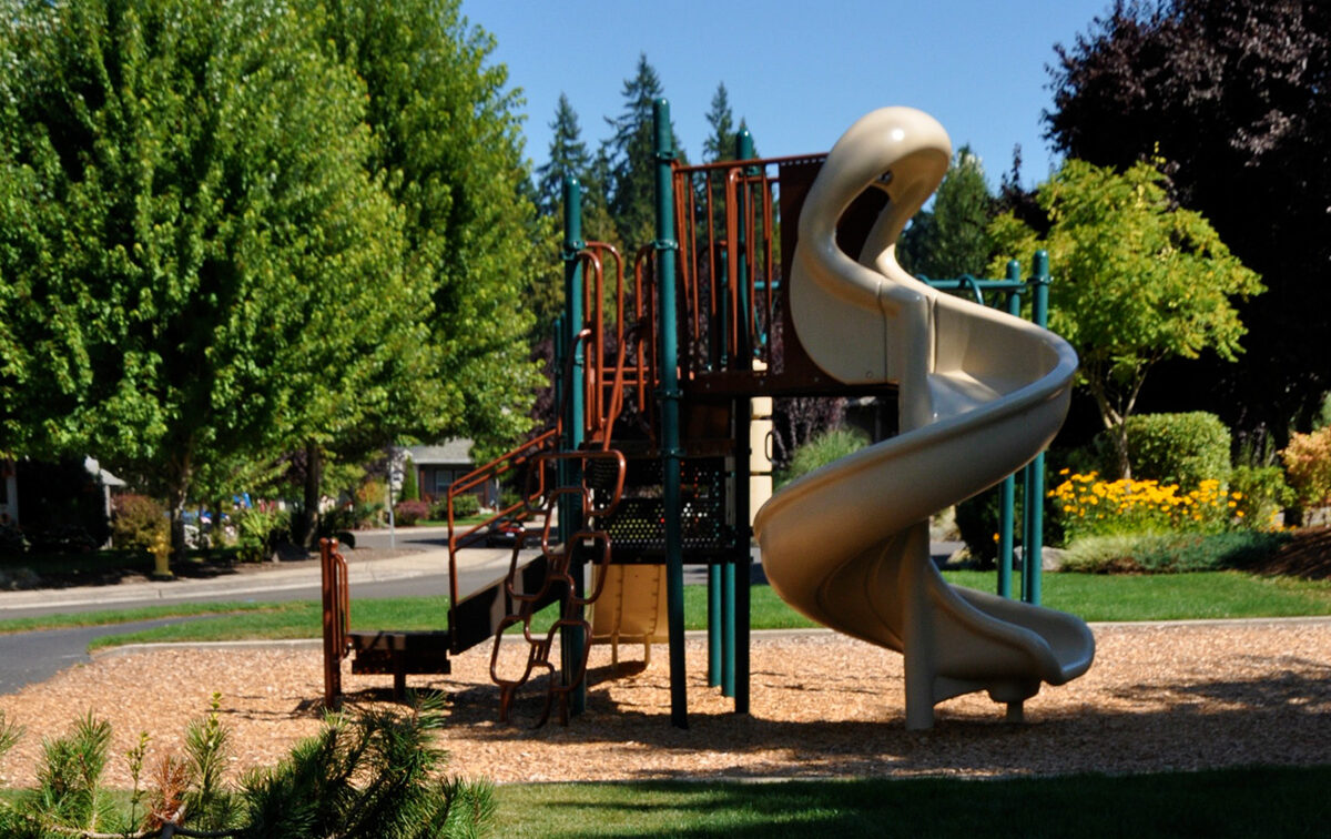 Playground with a spiral slide, climbing equipment, and wood chip ground covering, surrounded by trees and grass.