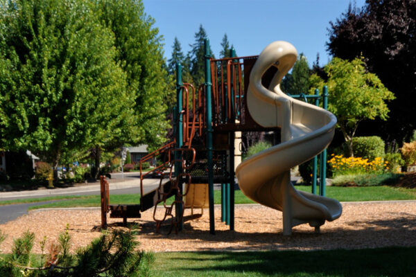 Playground with a spiral slide, climbing equipment, and wood chip ground covering, surrounded by trees and grass.