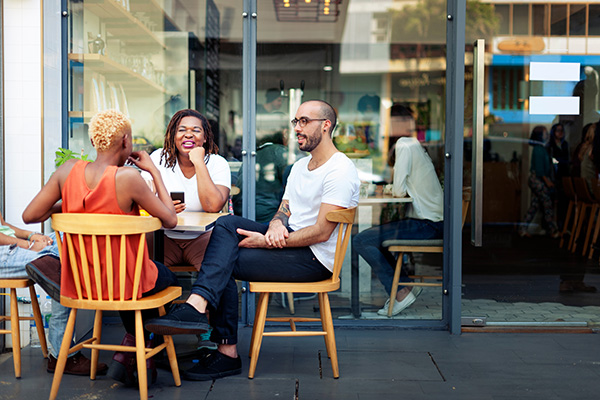 Four people sit at a table outside a cafe, engaged in conversation, with a glass window reflecting interior activity.