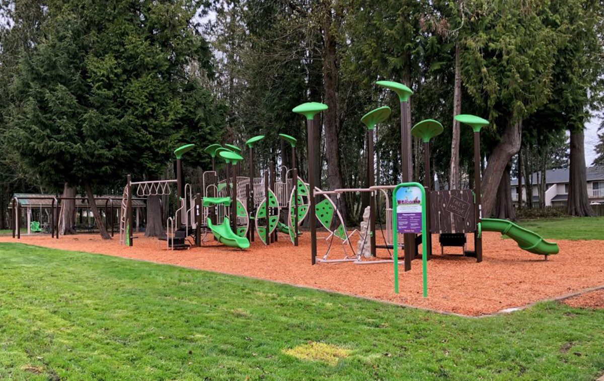 Children's playground with green slides, swings, climbing structures, and woodchip ground covering, surrounded by trees and grass.