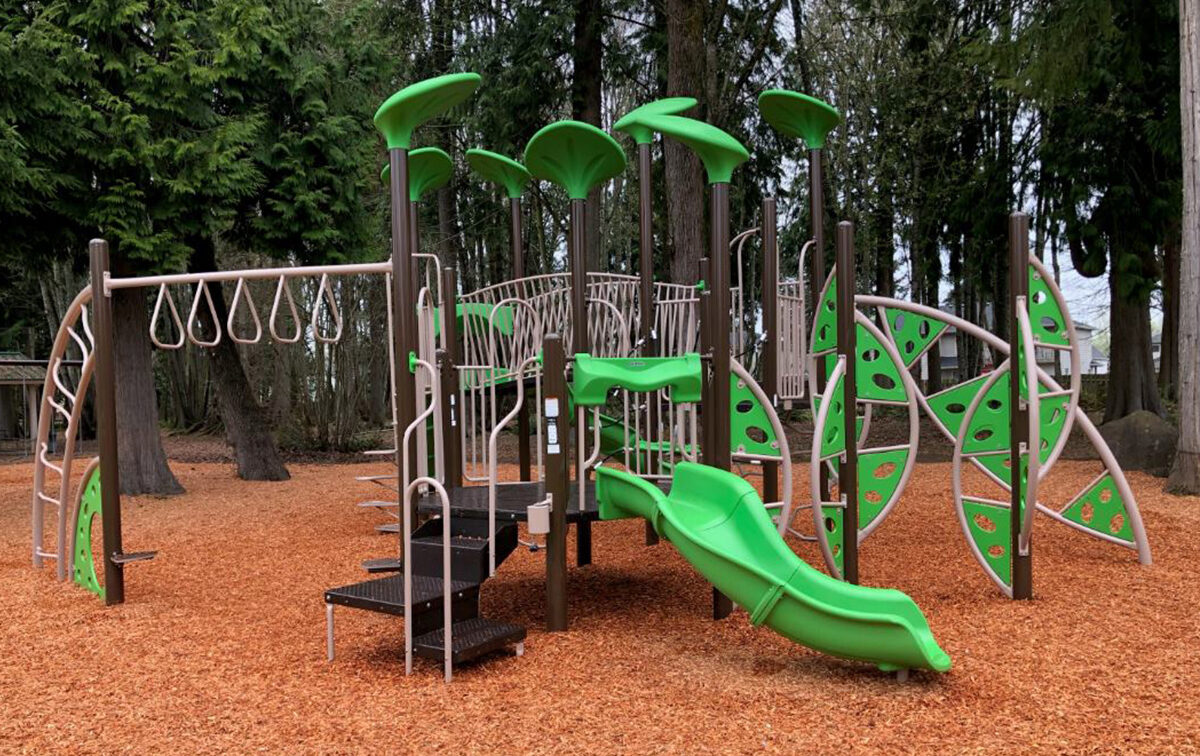 A children's playground set with green slides, climbing structures, and monkey bars, surrounded by wood chips and trees in the background.