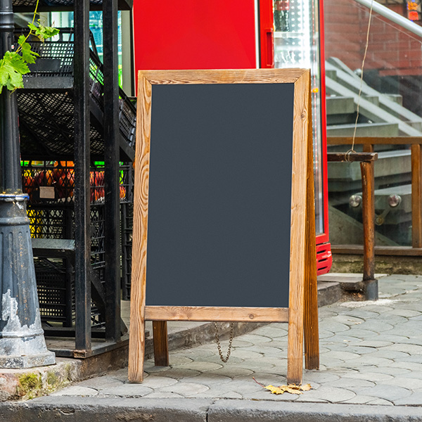 A blank chalkboard sign with a wooden frame stands on a sidewalk in front of a red vending machine and black crates.