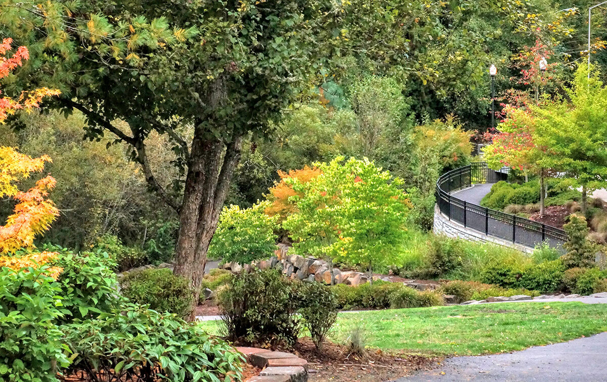 A curving paved path through a lush park with green and autumnal trees, bordered by a black metal fence and stone edging.