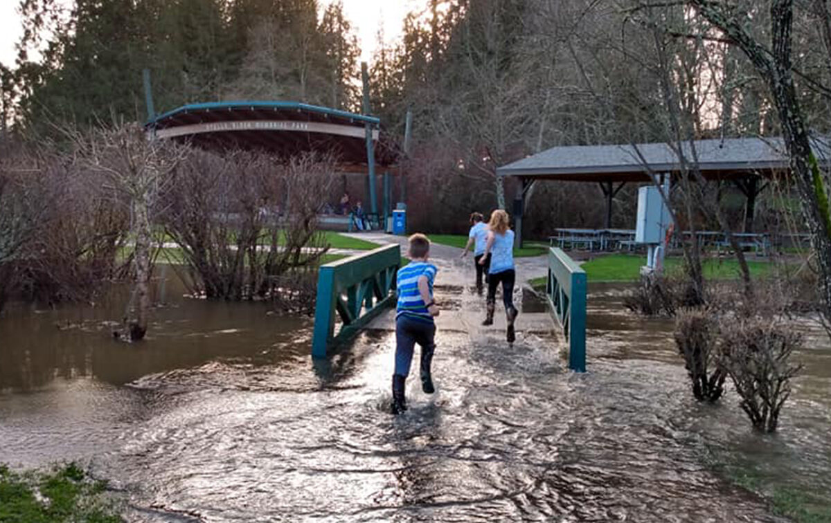 Two people run across a small bridge surrounded by floodwaters in a park with trees and structures in the background.