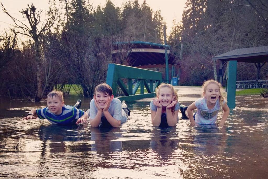 Four children lying on a flooded path smiling, surrounded by trees and a partially submerged bench, under a cloudy sky.