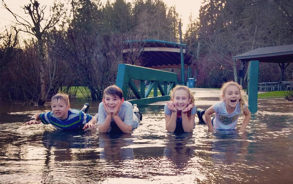 Four children lying on a flooded path smiling, surrounded by trees and a partially submerged bench, under a cloudy sky.