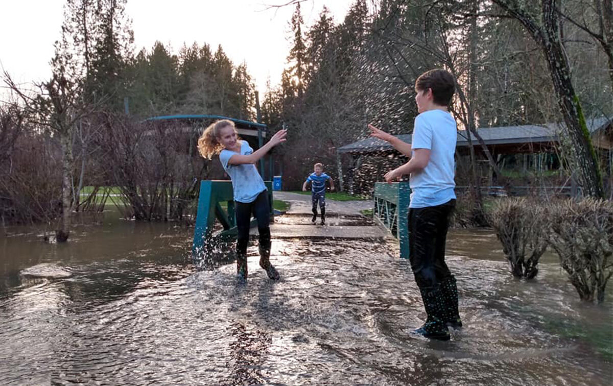 Two children play in shallow floodwater on a pathway, while another child approaches from a distance. Trees and a building are visible in the background.