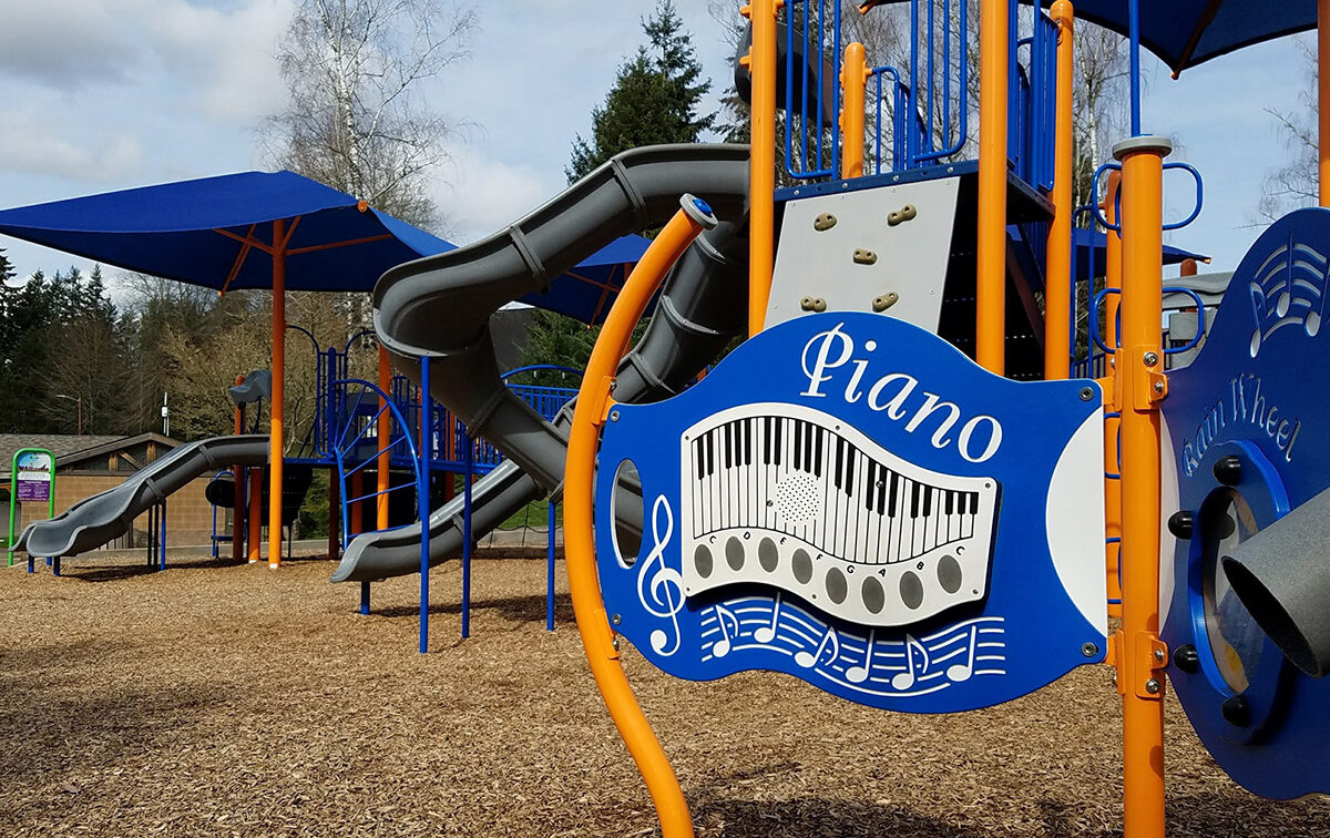 Playground featuring a blue and orange musical-themed structure with slides, a piano sign, and woodchip ground cover.