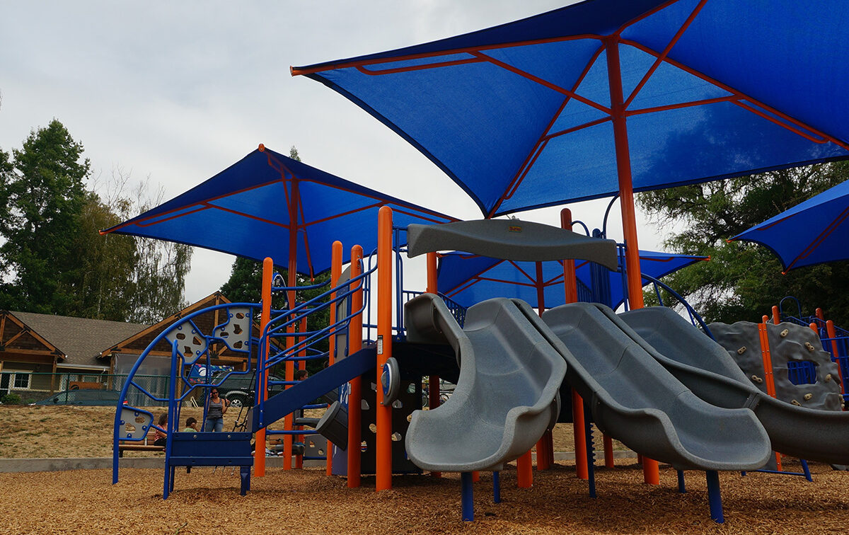 A playground with blue shade structures, featuring slides, climbing elements, and wood chip ground cover. Trees and a building are visible in the background under a cloudy sky.