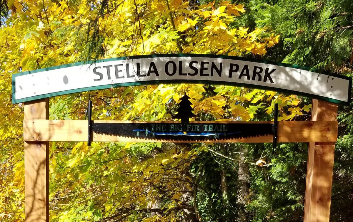 Entrance sign for Stella Olsen Park, featuring "The Big Fir Trail" text, surrounded by trees with autumn foliage.