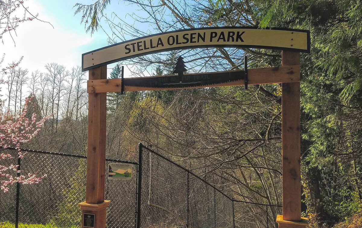 Entrance sign for Stella Olsen Park, framed by wooden posts, with a chain-link fence and trees in the background.