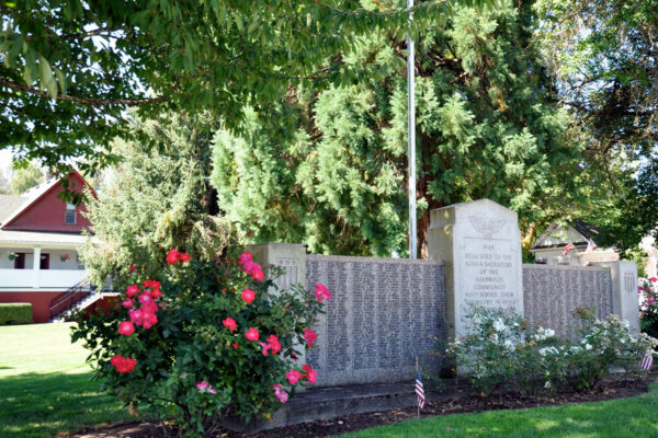 A memorial wall with engraved names stands under trees, surrounded by flowering bushes and small flags. A red-roofed house is visible in the background.