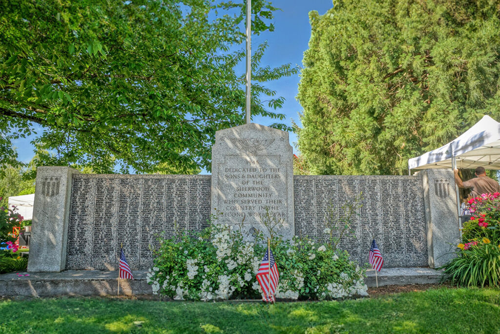 Memorial wall with engraved names, surrounded by greenery and American flags, honoring Sons and Daughters of Sherwood for military service.