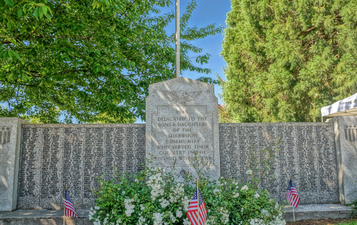 Stone war memorial with engraved text dedicated to community members who served, surrounded by white flowers, small American flags, and trees in the background.