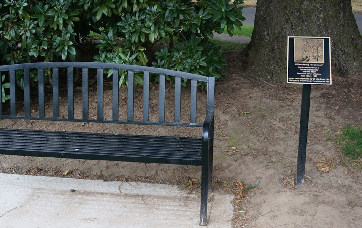 A black metal bench on a concrete surface next to a plaque mounted on a post. The bench is adjacent to a tree and surrounded by green foliage.