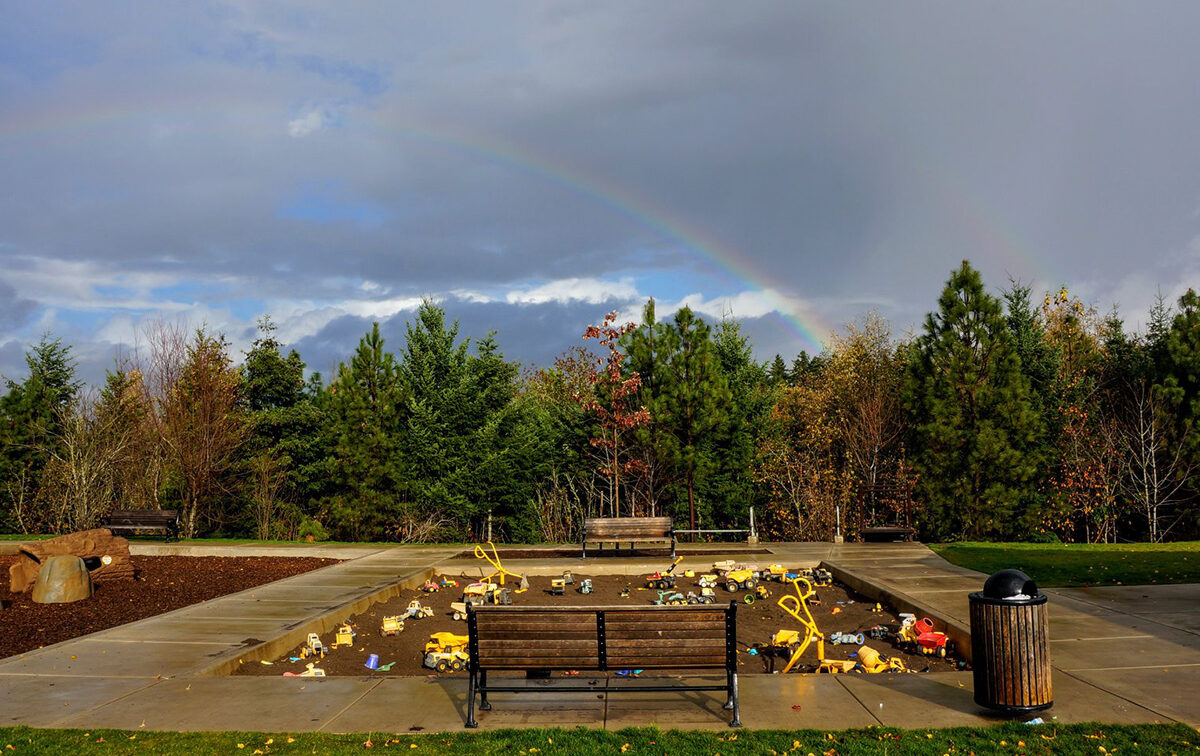 Children's playground with sandpit and toys, surrounded by trees and benches, under a cloudy sky with a rainbow.