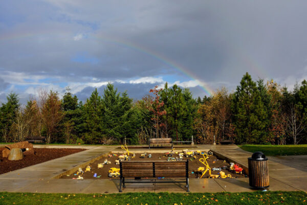 Children's playground with sandpit and toys, surrounded by trees and benches, under a cloudy sky with a rainbow.
