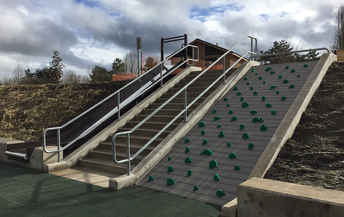 Playground area with a slide, stairs, and climbing wall with green holds under a cloudy sky.