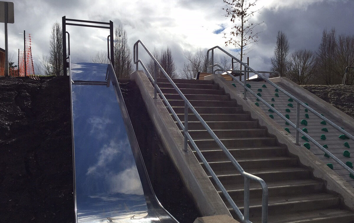 A metal slide is positioned next to a set of concrete stairs with handrails. The sky is cloudy, and trees are visible in the background.