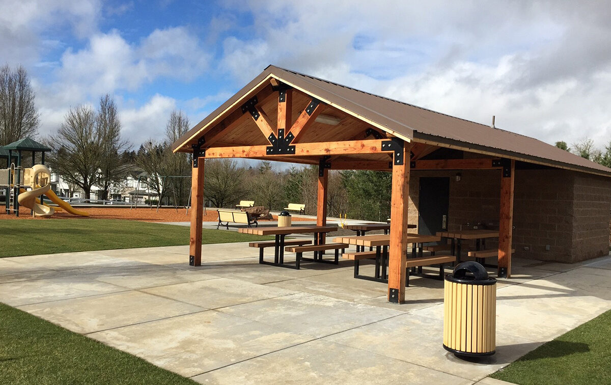 A park shelter with wooden beams, picnic tables, and a nearby trash can. A playground is visible in the background. Cloudy sky overhead.