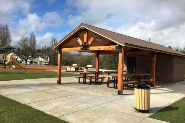A park shelter with wooden beams, picnic tables, and a nearby trash can. A playground is visible in the background. Cloudy sky overhead.
