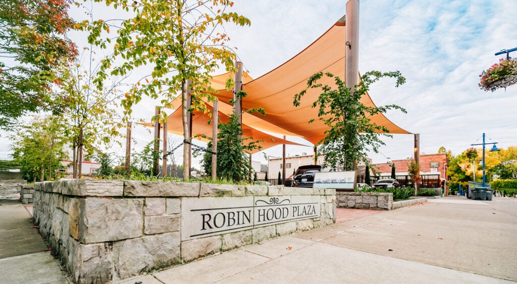 Outdoor plaza with orange canopy, stone wall, and young trees. Sign reads "Robin Hood Plaza." Buildings and cloudy sky in the background.