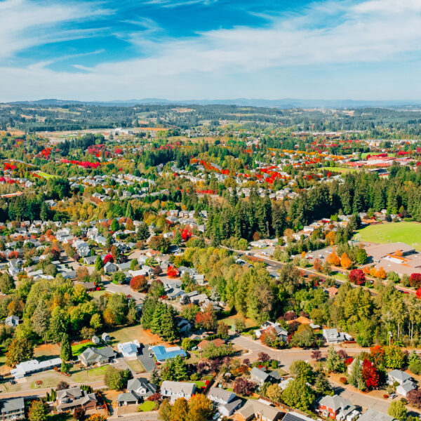 Aerial view of a suburban neighborhood surrounded by dense forests and open fields, featuring numerous homes and colorful autumn trees under a clear blue sky.