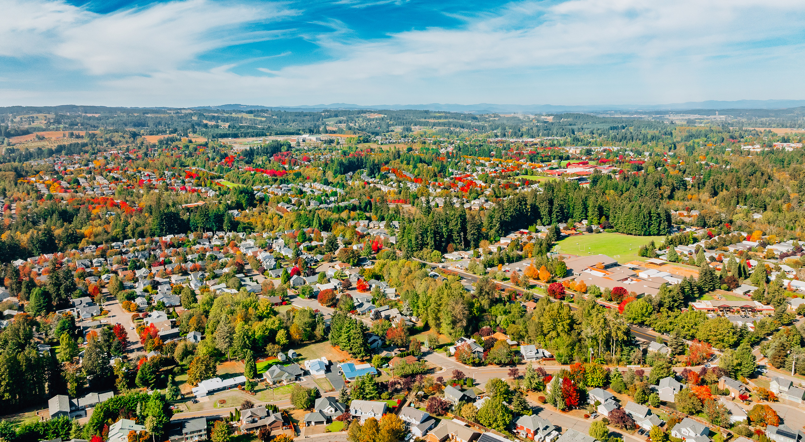 Aerial view of a suburban neighborhood surrounded by dense forests and open fields, featuring numerous homes and colorful autumn trees under a clear blue sky.