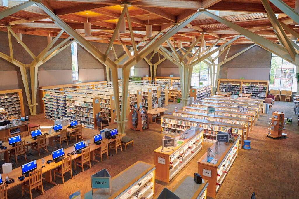 Spacious library interior with wooden ceiling beams, large windows, bookshelves, and several computer stations. Tables and chairs arranged for reading and study.