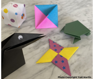 Origami creations including a black bird, green frog, yellow and pink star, multicolored box, and pink and blue pyramid on a marble surface.