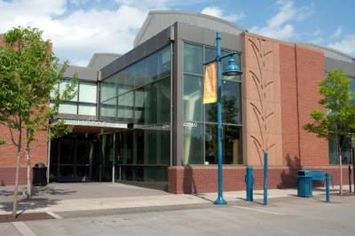 Modern brick and glass building with large windows, blue lamp posts, trees, and a sidewalk in front under a partly cloudy sky.